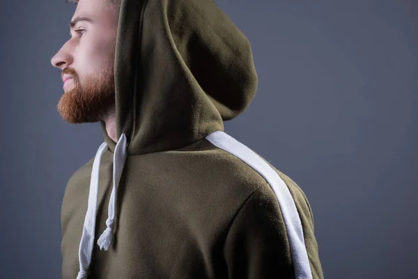 Studio portrait of a young bearded guy of twenty-five years old. in a sports jacket with a swamp-colored hood. On a gray background. Advertising photo of a handsome guy. in a sports warm jacket with a hood. green military color. In wireless headphone