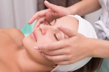 Kız masaj terapisti güzel bir kadınla yüz masajı yapıyor. Eller kapanıyor. Masaj odası. Spa yüz bakımı. Yüzün kasları için egzersizler. Rahatlatıcı yüz masajı