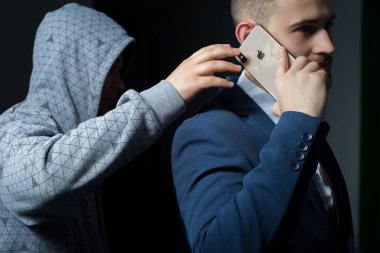 Zaporozhye. Ukraine. February 2020. Close-up portrait of a young bearded guy in a business suit holding an iPhone XS max. A robber in a dark hood, trying to steal a smartphone from behind. New smartphone from Apple. Modern technologies in business. B