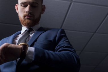İş elbisesi içinde genç sakallı yakışıklı bir adamın portresi, kameraya bakıyor, kol saatini gösteriyor, deri bir kayışla. Reklamcı erkek saatleri. Gri bir arka planda. İş adamı portresi