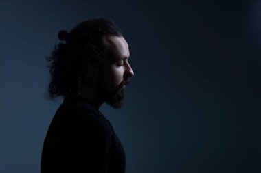 Otuz yaşında, uzun saçlı, sakallı bir gencin dramatik portresi aşağıya bakıyor. Karanlık bir arka planda. Yüzünde bir ışık huzmesi. Dramatik ışık, koyu erkek portresi, etkileyici adam, boş alan. Düşük tuşlu bir portre.
