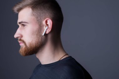 25 yaşında genç sakallı bir adamın portresi. Profilde duruyor. Kablosuz beyaz kulaklıkla. Sportif tarzda. Sutdiy, kulaklıklı sporcunun reklam fotoğrafı. Spor ve müzik. Adam kulaklıkla müzik dinliyor.