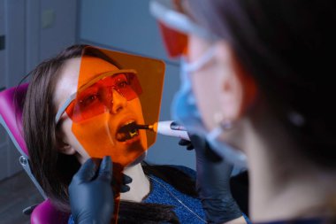Profesyonel diş beyazlatma prosedürünü uygulayan bir dişçi doktoru portresi ultraviyole radyasyon kullanıyor. Koruyucu turuncu gözlüklü hasta ve doktor. Gri bir arka planda. Dişçi kliniğinde. Hastalıkların önlenmesi ve tedavisi
