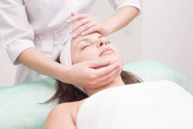 Kız masaj terapisti güzel bir kadınla yüz masajı yapıyor. Eller kapanıyor. Masaj odası. Spa yüz bakımı. Yüzün kasları için egzersizler. Rahatlatıcı yüz masajı