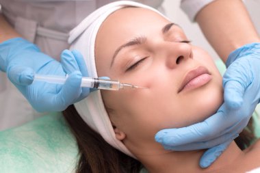 Genç bir kadının cildine enjekte edilmiş. Yakın plan. Tamam. İğneli bir şırıngayla güzellik uzmanı yüzündeki kırışıklıklara bir çare sunar. Kadınlarda yüz derisinin erken yaşlanma ile mücadele