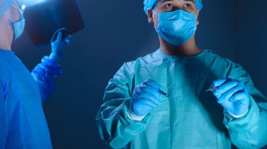 Steril elbiseli, şapkalı ve maskeli iki cerrahın portresi. Ameliyata hazırlanan bir cerrah elinde şırınga tutacağı olan bir neşter tutuyor, başka bir doktor da hastanın röntgenini çekip muayene ediyor. İşletim lambasının arka planında.