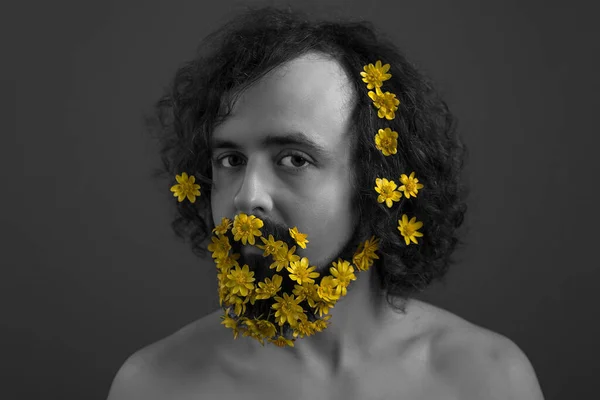 Studio black and white portrait of a young guy of thirty years old, yellow flowers woven into curly hair and a long beard. Concept of spring flowers in a beard. Flowers and spring. Brown-haired young guy with curly hair. Brutal bearded man with butte