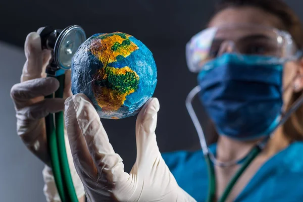 Closeup portrait of a female doctor in a protective mask, gloves, glasses, using a phonendoscope, auscultates the model of planet Earth. Studio photo on a gray background. Healing the whole planet. Pandemic, diseases that have swallowed the whole wor
