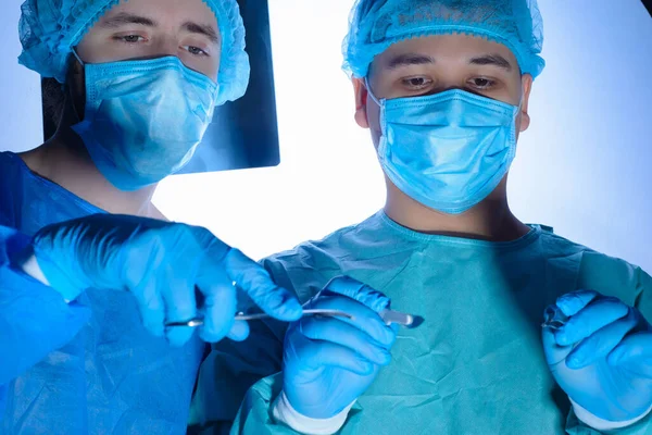 Portrait of two surgeons in sterile gowns, hats and masks. One surgeon, preparing to operate, holds a scalpel with a needle holder in his hands, another doctor assisting the first surgeon. Against the background of the operating lamp. Studio photo me