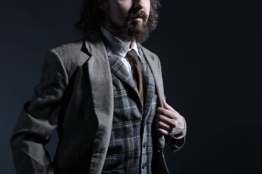 Otuz yıllık pahalı bir iş elbisesi içindeki sakallı ve kıvırcık saçlı genç bir adamın stüdyo draması elini ceketinin yakasından tutuyor. Karanlık bir arka planda. Yaratıcı insan portresi. Erkek ceketi reklamları, resmi takım elbiseler. Rembrandt Işığı