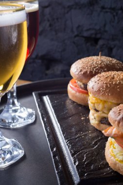 Hamburger ve patatesli bir kupa bira, el yapımı koyu ve köpüklü bira bardakları, hamburgerlerin arka planında, Shyomga, sığır eti köftesi, peynirli burger gibi çeşitli malzemelerle. Köy tarzında, bir siyaha karşı.