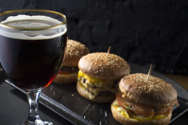 mug of beer with hamburger and fries close-up, beer glasses with craft dark and light beer with foam, against a background of burgers, with a variety of ingredients, including shyomga, beef meat patty, cheese burger. In a rustic style, against a blac