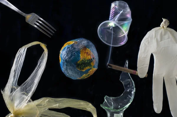 The concept of planet Earth pollution by plastic, metal, polyethylene, glass, rubber, smoke. Around the model of planet Earth, against the background of space, a rubber medical glove levitates, resembles a person, a steel fork, a plastic bag, a crump