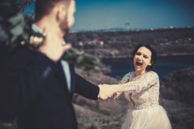 Düğün çiçekleriyle duran damadın elini tutan neşeli bir gelinin portresi. Açıl. Nehrin arka planına karşı. Düğün günü ve evlilik. Neşeli genç 25 güzel gelin ve damat doğal bir arka planda