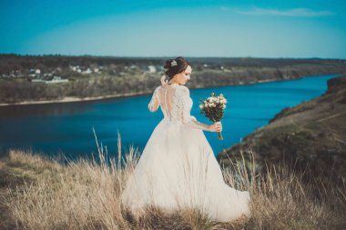 Genç bir gelinin sırtından genel plan portresi, elinde bir düğün buketi, bir nehirle manzaranın mesafesine bakıyor. Yaratıcı sanatsal boyama. 25 yıllık genç ve güzel bir gelinin düğün fotoğrafı. Tatili bekliyor.