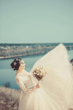 Genç bir gelinin sırtından genel plan portresi, elinde bir düğün buketi, bir nehirle manzaranın mesafesine bakıyor. Yaratıcı sanatsal boyama. 25 yıllık genç ve güzel bir gelinin düğün fotoğrafı. Tatili bekliyor.