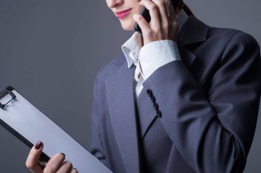 Yakın çekim stüdyo portresi, gri takım elbiseli iş kadını, ceket. Elinde boş bir kağıtla bir klasörü tutarak, akıllı bir telefonla konuşarak. Gri bir arka planda. Promosyon fotoğrafı. Ofis tarzının ticari fotoğrafı, önemli belgelerin imzalanması.