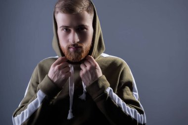 25 yaşında sakallı bir gencin stüdyo portresi. Bataklık renkli kapüşonlu bir spor ceketle. Gri bir arka planda. Yakışıklı bir adamın reklâm fotoğrafı. Kapüşonlu, spor ceketli. yeşil askeri renk. Kablosuz kulaklıkla.