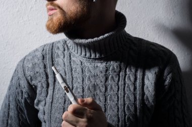 Yakın plan. Modern yüksek teknolojili elektronik sigara içen sakallı bir gencin sanat portresi. Boğazında gri bir kazak vardı. Gri bir arka planda. Stüdyo fotoğrafı. Sigara dumanı üflüyor. Modern sigara içmenin zararı. Iqos purosu.
