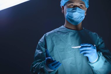 Steril takım elbiseli, maskeli, elinde neşter tutan ve ameliyata hazırlanan beyaz bir doktor cerrahın yakın plan portresi. Mavi arka planda. Ameliyat odası. Ameliyat mı? Üniformalı bir doktorun portresi ve koruyucu bir giysi. Yeniden yapılandırıcı ve 