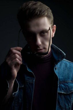 Genç bir adamın yakın plan portresi, kameraya bakıyor, gözlük takıyor. Denim tarzı. Gri bir arka planda. Y jenerasyonundaki bir adamın bin yıllık portresi. Dramatik bir portre, koyu renkli, gri arka planda erkek portresi. Kum ceketi, güneş gözlüğü.