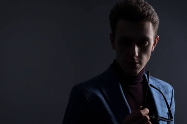 Genç bir adamın koyu renkli dramatik portresi, takım elbiseli bir iş adamı, gri bir arka planda başka yere bakıyor. Minimalist portre. Koyu renk erkek işi katı bir portre. Genç iş adamları. Reklam için boş yer. Erkek ceketi, kravat, p