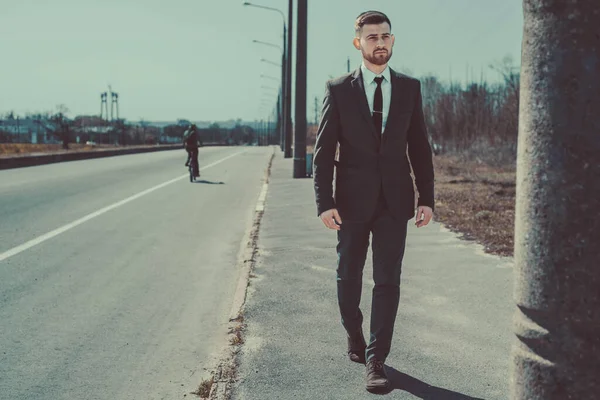 The lower torso of a man walking down the sidewalk along the side of the road. Portrait of a young bearded guy in a business suit walking along the road. Outdoor. Businessman by the road, walking, posing, looking to the side.