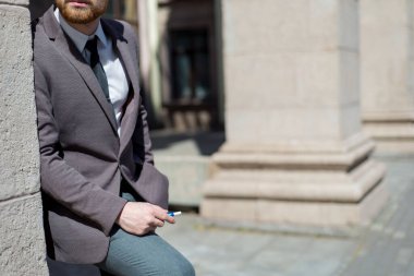 Elektronik sigara Iqos. 25 yaşında sakallı genç bir adamın portresi binanın dokulu duvarının yanında duruyor, sigara içiyor. Açık havada. Iqos sigarası kullanan yenilikçi yüksek teknoloji nikotin. Sigara reklamı. Sakal