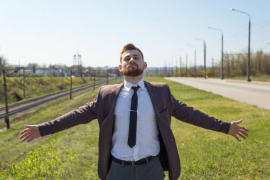 Genç bayan temiz havanın tadını çıkarıyor. 25 yaşındaki genç bir adamın portresi, iş elbisesi içinde, kolları açık, yol kenarında gündüz vakti boş bir trafiğin arka planında ve hareket hâlindeki demiryolu bulanık.