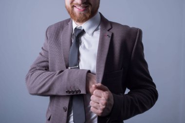 25 yaşında, resmi takım elbiseli, sakallı yakışıklı bir adamın stüdyo portresi elini ceketinin iç cebine sokuyor. Gri bir arka planda. Resmi tarzda. Ofis tarzı. Moda ve ofis