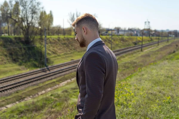 田舎道を歩いて若い男25歳の髭を生やした男の肖像画は ビジネススーツで 午後には空の道路に立っている 鉄道の背景にぼやけて — ストック写真