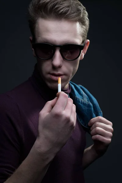手にタバコを持ちながらカメラを見てサングラスをかけた若い男の肖像画を閉じ タバコを灯す準備をしていた デニムスタイル 灰色の背景で ミレニアム世代の男の肖像画よ 劇的な肖像画 — ストック写真