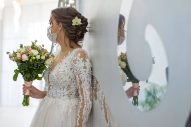 Coronavirus salgını sırasında şehrin caddesinde maske takan gelin. Düğün günü COVID-19 koruma düğünü ve Coronavirus covid 19-20. Koruyucu bir tıbbi maske takmış, gelinlik giymiş gelin elinde bir buket düğün buketi ile bulanık bir şekilde dikiliyor., 