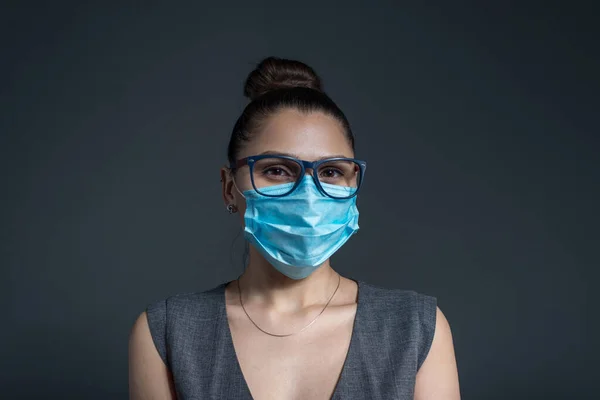 Girl in a protective medical mask looking at the camera. Studio photo on a gray background. Work in the mask. Office work during quarantine in a medical protective mask. Business style gray dress, a girl stands with a folder for papers. Covid 19-20.