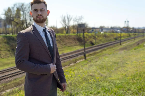 田舎道を歩いて若い男25歳の髭を生やした男の肖像画は ビジネススーツで 午後には空の道路に立っている 鉄道の背景にぼやけて — ストック写真