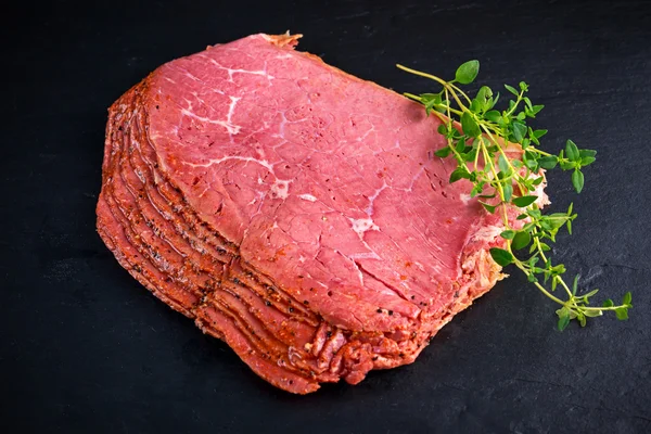 Rodajas de pastrami de roast beef con pimienta sobre fondo de piedra negra con hierbas —  Fotos de Stock