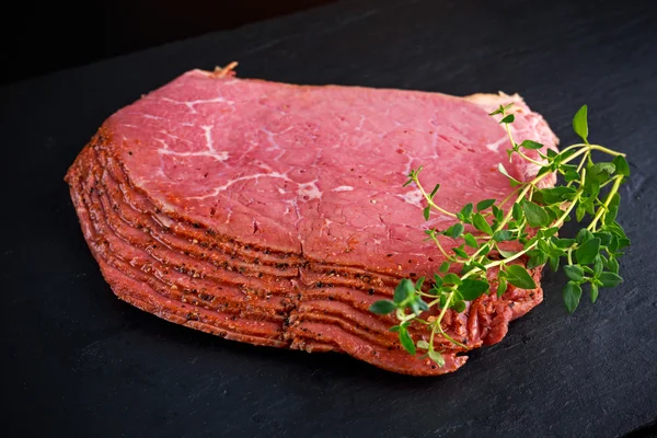 Peppered roast beef pastrami slices no fundo de pedra preta com ervas — Fotografia de Stock