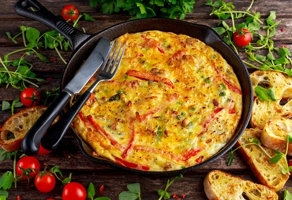 Frittata gemaakt van eieren, aardappel, spek, paprika, peterselie, groene erwten, ui, kaas in IJzeren pan. op houten tafel. — Stockfoto