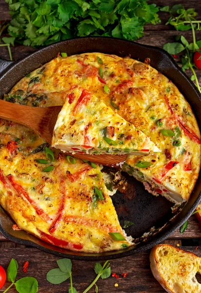 Frittata gemaakt van eieren, aardappel, spek, paprika, peterselie, groene erwten, ui, kaas in IJzeren pan. op houten tafel. — Stockfoto