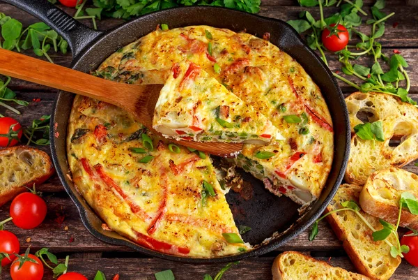 Frittata gemaakt van eieren, aardappel, spek, paprika, peterselie, groene erwten, ui, kaas in IJzeren pan. op houten tafel. — Stockfoto