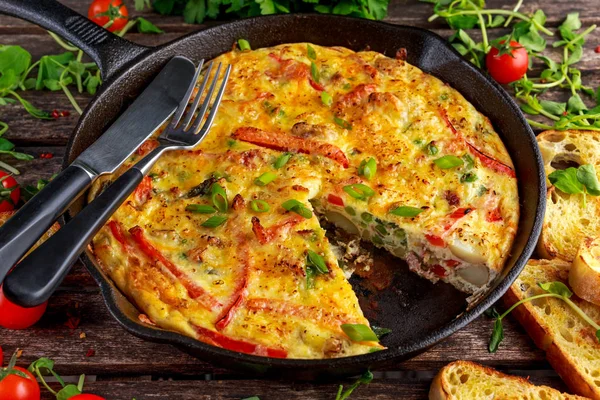 Frittata gemaakt van eieren, aardappel, spek, paprika, peterselie, groene erwten, ui, kaas in IJzeren pan. op houten tafel. — Stockfoto