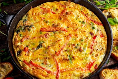 Yumurta, patates, pastırma, kırmızı biber, maydanoz, yeşil bezelye, soğan, demir tavada peynir Frittata yapılmış. ahşap masa.