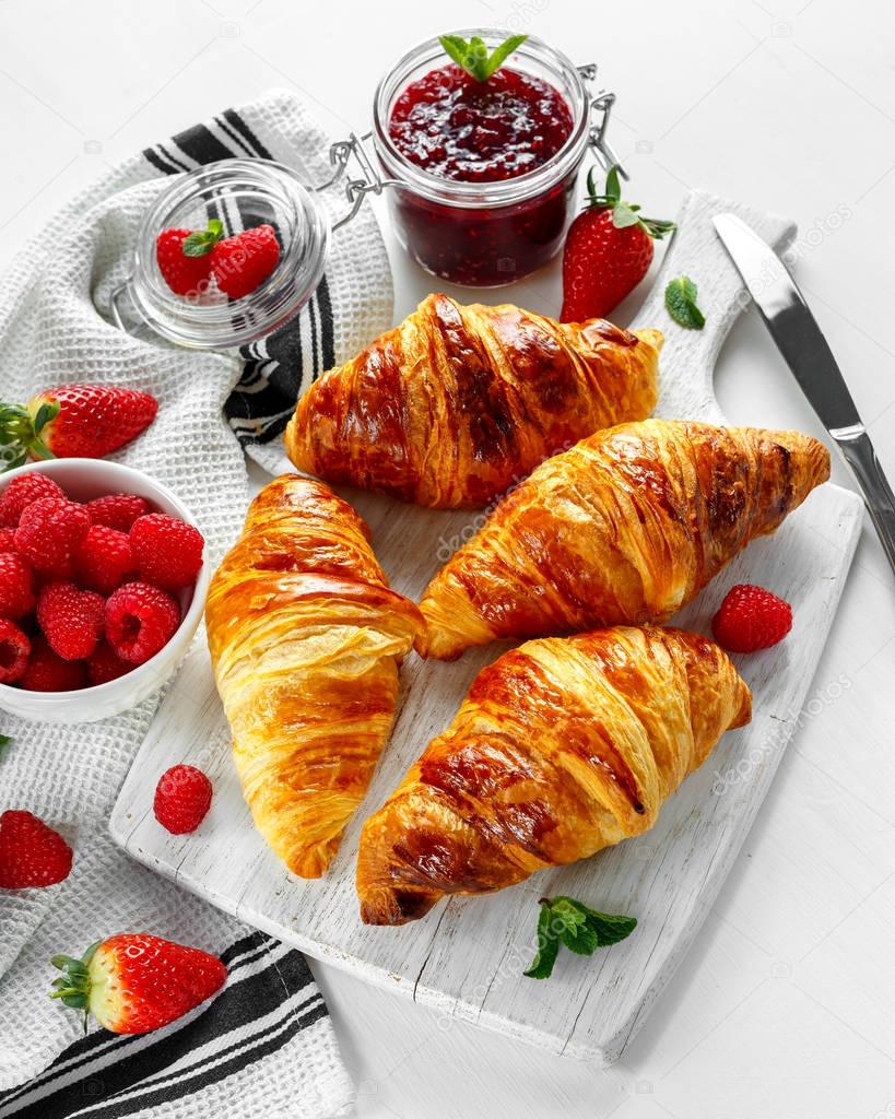 Fresh tasty homemade croissants with ripe berries and raspberries jam on white wooden board