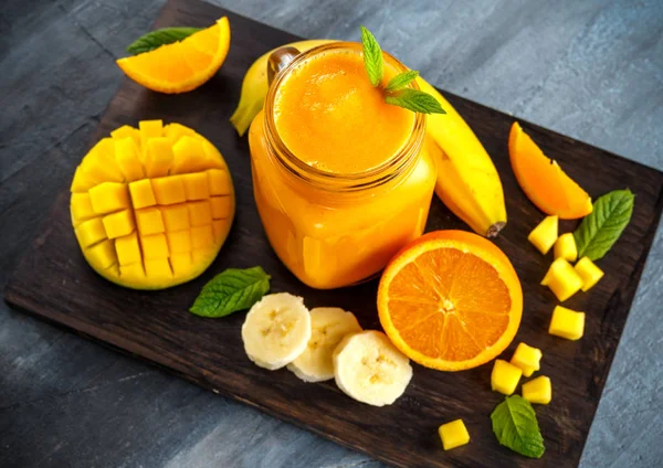 Bebida de batido de naranja fresca con plátano, mango, zanahorias en tablero de madera negro . — Foto de Stock