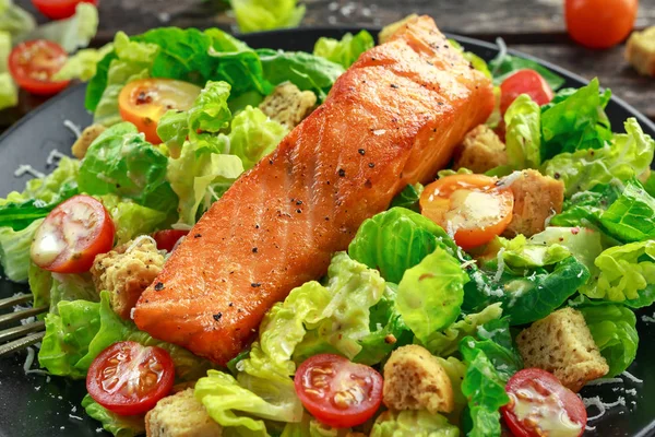 Caesar Salmon Salad with croutons, parmesan cheese, tomatoes, dressing and pepper in a black plate on wooden table. — Stock Photo, Image