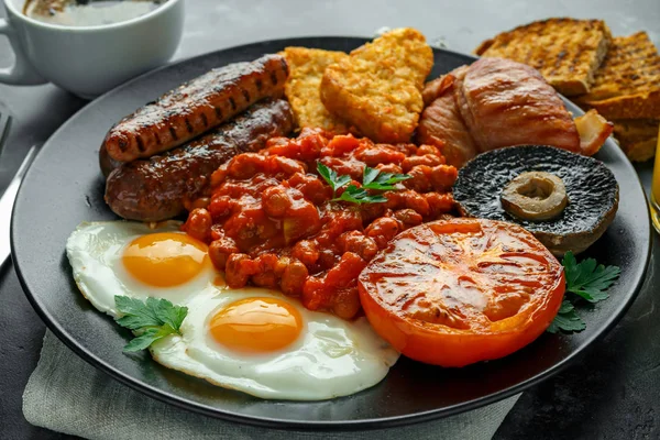 Café da manhã inglês completo com bacon, salsicha, ovo frito, feijão assado, batatas fritas e cogumelos em prato preto. copo de café . — Fotografia de Stock