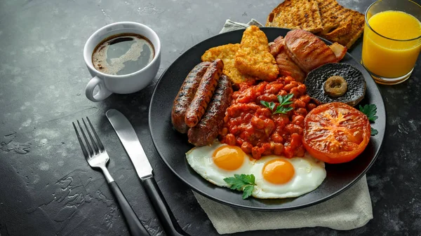 Full English breakfast with bacon, sausage, fried egg, baked beans, hash browns and mushrooms in black plate. cup coffe, orange juice — Stock Photo, Image