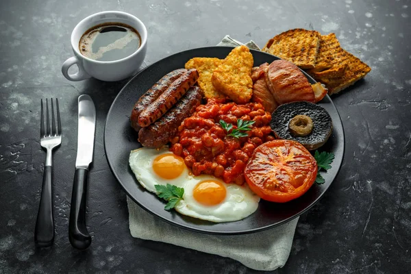Petit déjeuner anglais complet avec bacon, saucisse, oeuf frit, haricots cuits au four, pommes de terre hachées et champignons dans une assiette noire. tasse de café . — Photo