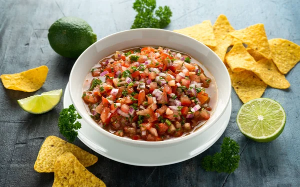 Мексиканский томатный сальса в белой миске с лаймом, красный лук, перец халапеньо, петрушка и чипсы с тортильей — стоковое фото