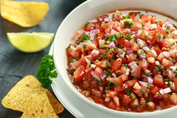 Mexican Tomato Salsa in white bowl with lime, red onion, jalapeno pepper, parsley and tortilla chips — Stock Photo, Image